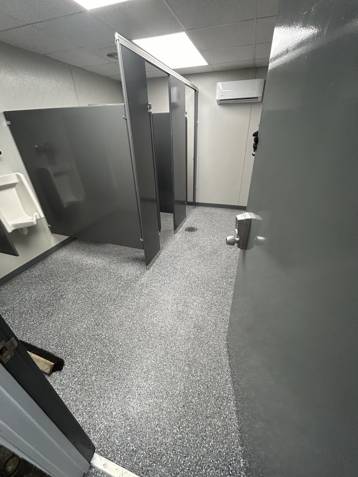 Completed commercial restroom with durable speckled flooring, partitions, urinals, and clean gray finishes